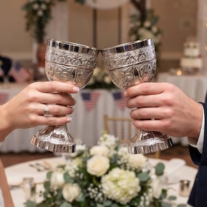 May include: Two silver-toned metal goblets with intricate embossed floral and geometric patterns on the bowls and stems. The goblets are held up in a toasting gesture, showcasing the metallic finish and detailed craftsmanship of the vessels.