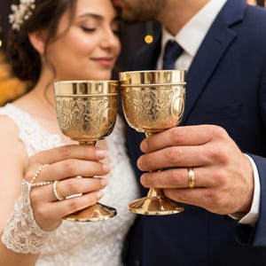 May include: Two matching gold-toned metal goblets featuring intricate embossed floral and scrollwork patterns around the bowl. Each chalice has a smooth, polished rim and a sturdy stem, designed with a classic silhouette for ceremonial use.