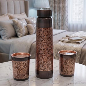 May include: A copper water bottle and two matching cups sit on a marble table. The bottle and cups have a dark brown base with an ornate, repeating copper-colored floral pattern. The bottle has a dark brown lid.
