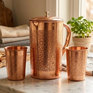 May include: A copper pitcher with a lid and handle, accompanied by two matching copper tumblers. All pieces feature an embossed floral pattern. The set is displayed on a marble countertop, suggesting a kitchen or dining setting.