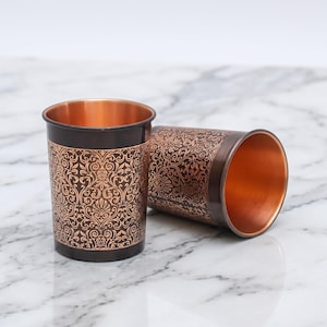 May include: Two copper-colored metal drinking glasses. One is upright, the other is lying on its side. Both have a dark brown exterior with an ornate, floral pattern and a bright copper interior.