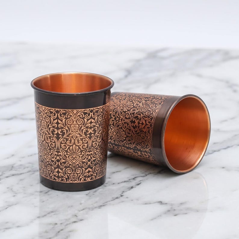 May include: Two copper-colored metal drinking glasses. One is upright, the other is lying on its side. Both have a dark brown exterior with an ornate, floral pattern and a bright copper interior.