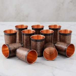 May include: A collection of copper-colored drinking glasses. The glasses have a dark brown band with an ornate floral pattern around the top. The interiors of the glasses are a bright copper color. Some glasses are upright, while others are lying on their sides.