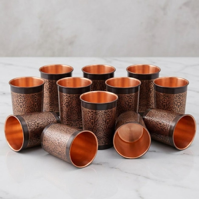 May include: A collection of copper-colored drinking glasses. The glasses have a dark brown band with an ornate floral pattern around the top. The interiors of the glasses are a bright copper color. Some glasses are upright, while others are lying on their sides.