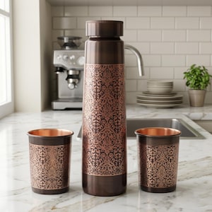 May include: A copper-colored water bottle and two matching tumblers. The bottle has a decorative floral pattern and a screw-on cap. The tumblers have the same pattern. The set is displayed on a marble countertop in a kitchen setting.