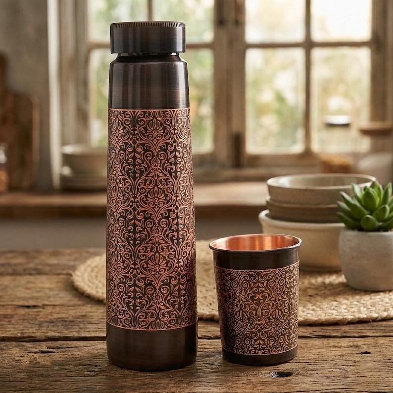 May include: A copper-colored water bottle and matching cup with an ornate, repeating floral pattern. The bottle has a dark brown cap and base. The cup is cylindrical. Both items are on a wooden surface.