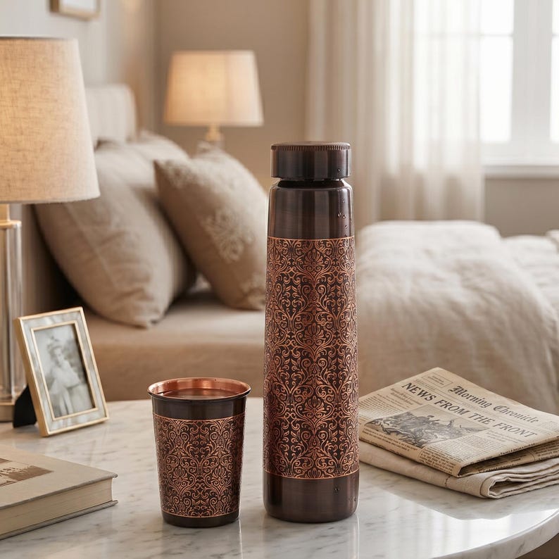 May include: A copper-colored water bottle and matching tumbler with an ornate, repeating floral pattern. The bottle has a dark brown cap and base. The tumbler is cylindrical and sits next to the bottle on a marble surface.