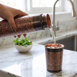 May include: A copper water bottle and cup with an ornate floral design. Water is being poured from the bottle into the cup. A small bowl of green herbs sits nearby on a marble countertop. The bottle and cup are made of copper.