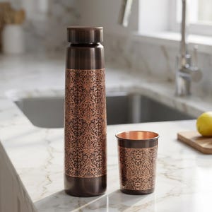 May include: A copper-colored water bottle and matching cup with an ornate, repeating floral pattern. The bottle has a screw-on cap. The cup is cylindrical. Both items are on a white countertop.