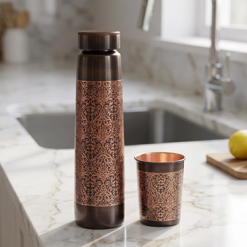 May include: A copper-colored water bottle and matching cup with an ornate, repeating floral pattern. The bottle has a screw-on cap. The cup is cylindrical. Both items are on a white countertop.