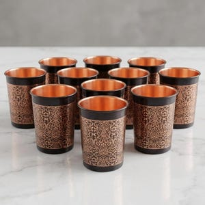 May include: A set of ten copper-colored drinking glasses with a black band and ornate floral pattern. The interior of the glasses is a bright copper color. The glasses are arranged on a white marble surface.
