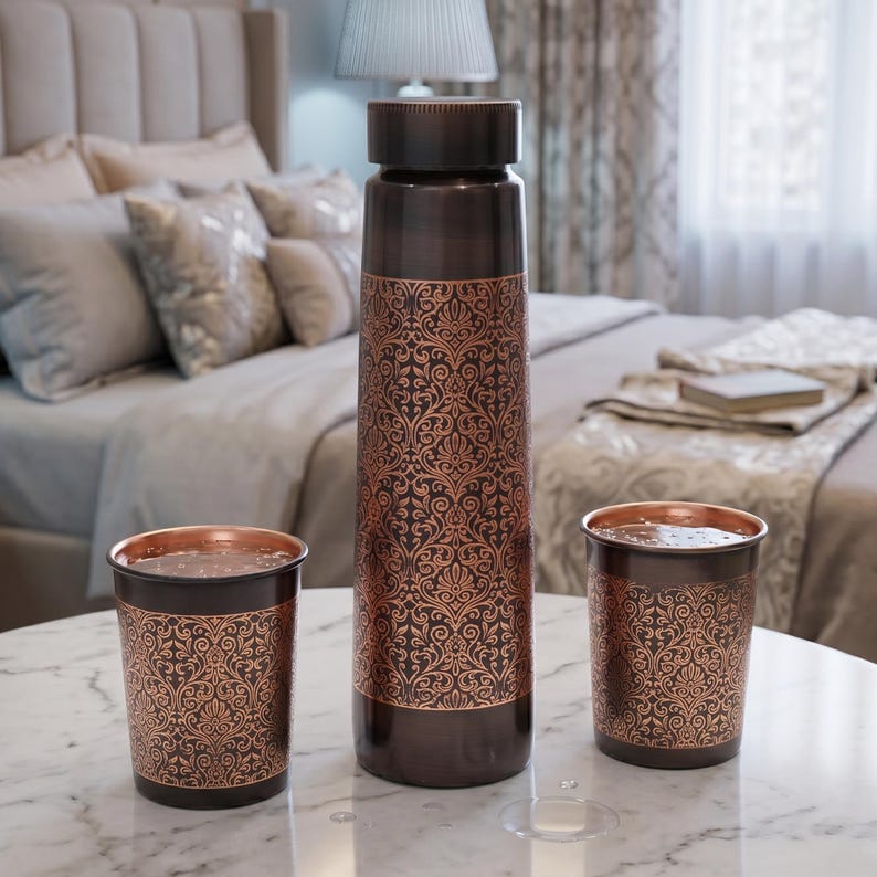 May include: A copper water bottle and two matching cups sit on a marble table. The bottle and cups have a dark brown base with an ornate, repeating copper-colored floral pattern. The bottle has a dark brown lid.