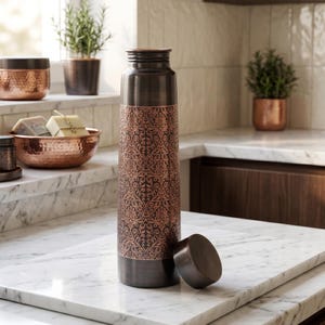 May include: A copper-colored water bottle with an intricate floral pattern. The bottle is cylindrical with a screw-on cap. The design is a mix of copper and dark bronze, with a matching cap. The bottle is sitting on a marble countertop.