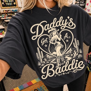 Peut inclure: Sweat-shirt gris foncé avec un motif crème. Le motif représente une illustration de cowgirl dans un cœur en forme de corde, avec les mots "Daddy's Got A Baddie". Le sweat-shirt a des manches courtes.