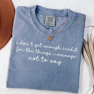 Pode incluir: Uma camiseta azul Comfort Colors tingida com o texto "i don't get enough credit for the things i manage not to say" em branco. Um colar de prata com um pingente redondo está sobre a camiseta. Tamanho M.
