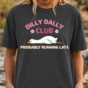 May include: A charcoal gray t-shirt featuring a graphic of a white goose lying down. The text reads DILLY DALLY CLUB in pink and PROBABLY RUNNING LATE in cream, with two small white flowers flanking the word CLUB on the front of the garment.