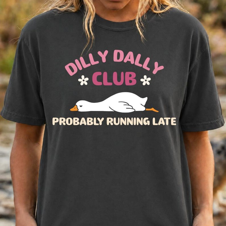 May include: A charcoal gray t-shirt featuring a graphic of a white goose lying down. The text reads DILLY DALLY CLUB in pink and PROBABLY RUNNING LATE in cream, with two small white flowers flanking the word CLUB on the front of the garment.
