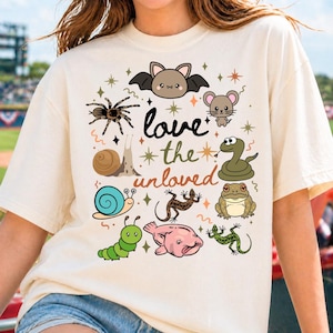 May include: Cream-colored t-shirt featuring a collection of illustrated animals, including a bat, mouse, spider, snail, snake, frog, lizard, caterpillar, and blobfish. The text "love the unloved" is written in a whimsical font.
