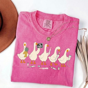 Puede incluir: Una camiseta rosa de manga corta con un gráfico de cinco gansos blancos. Los gansos sostienen una piña, una ecografía, una prueba de embarazo, una jeringa y un dispositivo médico. Debajo hay pequeños viales médicos y jeringas dispersos.