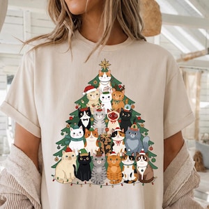 以下が含まれることがあります： ベージュの半袖Tシャツで、多数の猫のイラストで構成されたクリスマスツリーのグラフィックプリントが施されています。猫はさまざまな色や模様で描かれており、多くはクリスマスの帽子、スカーフ、リボンを身に着けています。