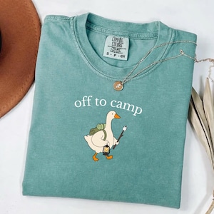 May include: A sage green short-sleeve t-shirt featuring a graphic of a white goose carrying a green backpack, a lantern, and a stick with a marshmallow. The text "off to camp" is printed above the goose in white. The shirt has a crew neckline.