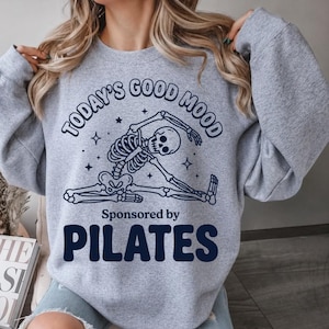 Può includere: Felpa grigio chiaro con il testo blu navy "TODAY'S GOOD MOOD" e "Sponsored by PILATES". Un'immagine di uno scheletro che si allunga. La felpa è uno stile casual e confortevole.