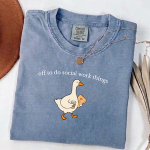 May include: A blue short-sleeved t-shirt with a crew neckline and a Comfort Colors tag indicating size M. The shirt features a graphic of a white goose holding a coffee cup and a tan folder with a heart, and the text off to do social work things.