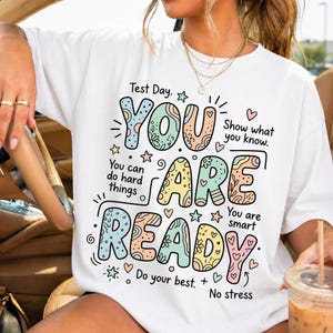 Puede incluir: Camiseta blanca con un gráfico colorido que dice "YOU ARE READY" en una fuente lúdica. El diseño incluye estrellas, corazones y los textos "Test Day", "You can do hard things" y "Show what you know."