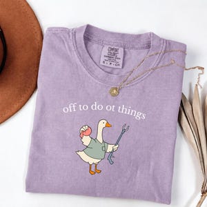 May include: A light purple t-shirt featuring a graphic of a white goose wearing a green shirt, holding a pink ball and a reacher grabber tool. The text "off to do ot things" is printed above the goose. The shirt has a Comfort Colors label.