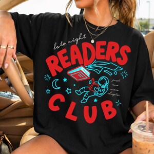 Op de afbeelding: Zwart T-shirt met een rood en blauw grafisch ontwerp. Het ontwerp bevat de woorden "late night READERS CLUB" en een astronaut die een boek leest. Sterren, een maan en de zin "just one more chapter" zijn ook aanwezig.