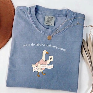 May include: A blue short-sleeved t-shirt with a graphic of a goose wearing a pink shirt, holding a card with footprints and a heart rhythm strip. The text reads, "off to do labor & delivery things." The shirt has a Comfort Colors size M label.