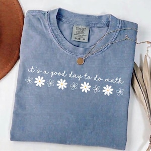 Puede incluir: Una camiseta azul Comfort Colors con la frase "it's a good day to do math" en escritura blanca y gráficos de margaritas. Un collar plateado con un colgante circular está sobre la camiseta. Talla M.