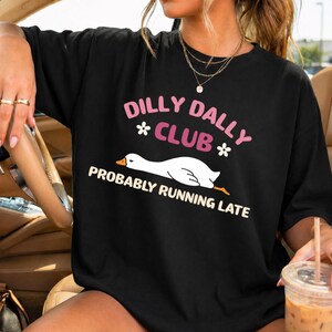May include: A black short-sleeve t-shirt featuring a graphic of a white goose and the text DILLY DALLY CLUB PROBABLY RUNNING LATE in pink and cream colors. The design includes two small white flower icons flanking the word CLUB.