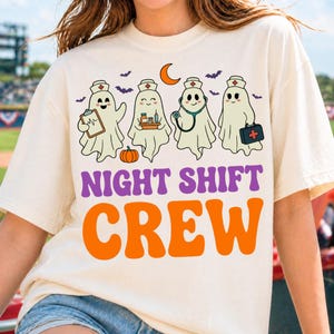 May include: A cream-colored t-shirt featuring a graphic of four ghosts dressed as nurses. The ghosts hold a clipboard, medicine, a stethoscope, and a medical bag. Below the ghosts, the text reads NIGHT SHIFT CREW in purple and orange letters.