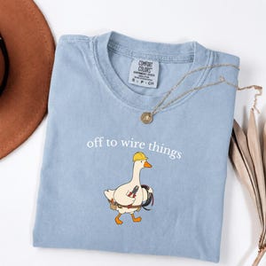 May include: A light blue t-shirt featuring a graphic of a white goose wearing a yellow hard hat, carrying a tool belt and a coil of black wire. The text "off to wire things" is printed above the goose. The shirt has a Comfort Colors label.