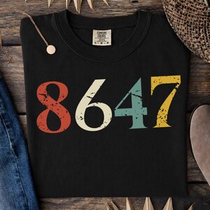 Może przedstawiać: Czarny t-shirt z okrągłym dekoltem z numerem "8647" w stylu retro. Numery są w odcieniach czerwieni, kremu, turkusu i żółci. Koszulka ma metkę Comfort Colors.