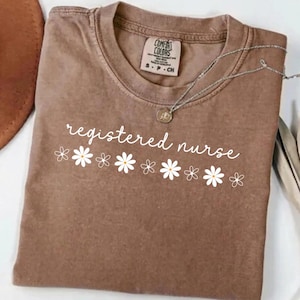 May include: A brown t-shirt with the words "registered nurse" in white script, decorated with white daisy graphics. A silver necklace with a pendant is draped across the shirt. The shirt has a Comfort Colors tag.