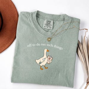 Può includere: Una t-shirt verde salvia con la grafica di un'oca bianca che tiene in mano una siringa e una cartellina con l'impronta di una zampa. Il testo "off to do vet tech things" è stampato sopra l'oca. La maglietta ha un girocollo e un'etichetta.