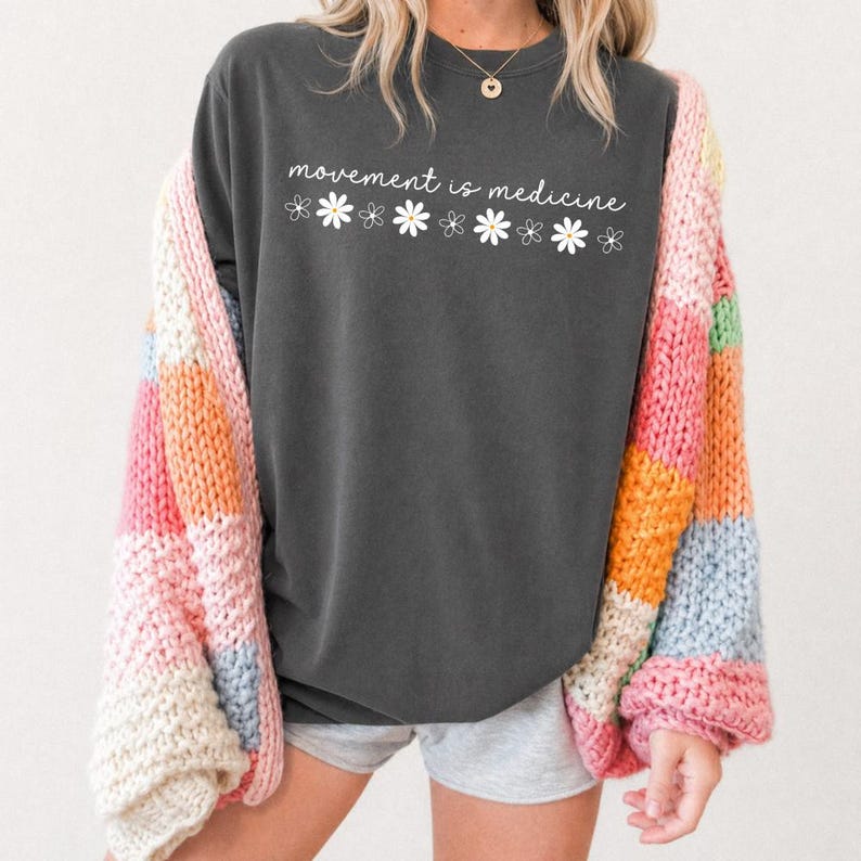 Puede incluir: Una camiseta de manga larga gris oscuro con el texto "movement is medicine" en escritura blanca, decorada con gr&aacute;ficos de margaritas blancas. Un c&aacute;rdigan de patchwork colorido en rosa, naranja, amarillo y azul se usa sobre la camisa.