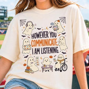 Op de afbeelding: Een crèmekleurig t-shirt met een afbeelding van zes cartoongeesten die verschillende communicatiemethoden gebruiken. De tekst luidt "HOWEVER YOU COMMUNICATE, I AM LISTENING". Het ontwerp bevat kleine pompoenen, vleermuizen en snoepjes.