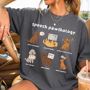 May include: Dark gray t-shirt with the text "Speech Pawthology" and illustrations of dogs. The shirt features various dog-related puns and diagrams, including "bark-nunciation" and "fur-nemes". A fun and educational design.