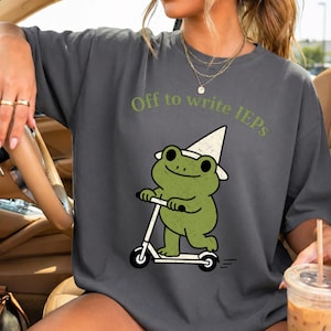 Könnte beinhalten: Dunkelgraues T-Shirt mit dem grünen Text "Off to write IEPs". Das Shirt zeigt einen Cartoon-Frosch mit weißem Hut, der einen weißen Roller fährt. Der Frosch ist grün mit schwarzen Augen und einem lächelnden Gesicht.