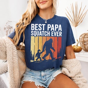 Può includere: T-shirt blu navy con la scritta bianca "BEST PAPA SQUATCH EVER". La grafica presenta una silhouette di Bigfoot che tiene per mano un bambino, su uno sfondo retrò a tramonto. Un capo d'abbigliamento comodo e casual.