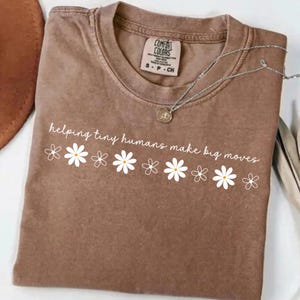 May include: A brown Comfort Colors t-shirt with the phrase "helping tiny humans make big moves" in white script. White daisy graphics are below the text. A silver necklace with a pendant is on the shirt.