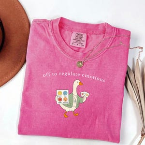 Puede incluir: Una camiseta rosa de manga corta con el gráfico de un ganso que lleva una rebeca verde y sostiene una tabla de emociones. El texto "off to regulate emotions" está impreso sobre el ganso. La camiseta tiene una etiqueta de la marca Comfort Colors.