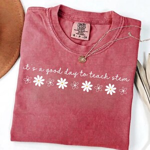May include: A dusty rose short-sleeve t-shirt featuring the white text "it's a good day to teach stem" above a horizontal row of white daisy illustrations. The shirt has a crew neckline and a Comfort Colors brand label at the inner collar.