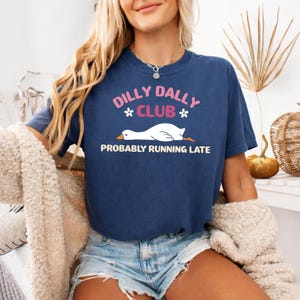 May include: A navy blue short-sleeve t-shirt featuring a graphic of a white goose lying down. The text reads DILLY DALLY CLUB in pink and PROBABLY RUNNING LATE in white, with two small white flowers flanking the word CLUB on the front of the shirt.