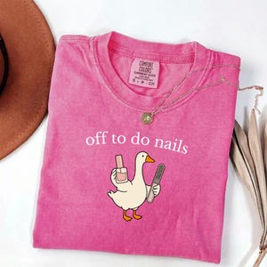 May include: A pink short-sleeved t-shirt featuring a graphic of a white goose holding a bottle of nail polish and a nail file. The text "off to do nails" is printed in white above the goose. The shirt has a Comfort Colors label at the neck.