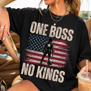 Può includere: T-shirt nero con un design a bandiera americana invecchiata e la scritta "ONE BOSS NO KINGS". La grafica include una figura che tiene una chitarra. Un capo d'abbigliamento casual.