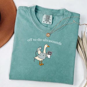 Op de afbeelding: Een saliegroen t-shirt met een afbeelding van een witte gans in een grijze tuniek, die een echoprobe en een echobeeld vasthoudt. De tekst "off to do ultrasounds" staat in witte kleine letters boven de gans gedrukt.